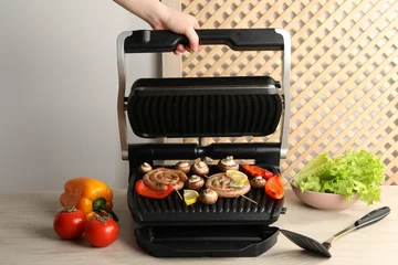  Woman cooking homemade sausages with bell peppers and mushrooms on electric grill at wooden table, closeup © New Africa