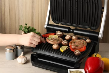  Woman cooking homemade sausages with mushrooms and bell pepper on electric grill at wooden table, closeup © New Africa