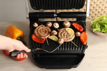 Woman cooking homemade sausages with bell peppers and mushrooms on electric grill at wooden table, closeup © New Africa