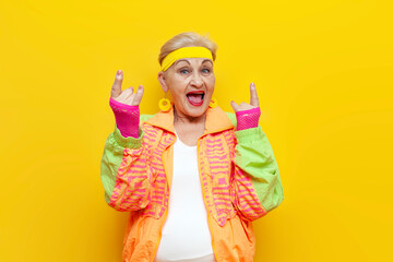 crazy funny old granny in sports colorful clothes screams and shows a rock gesture on a yellow...