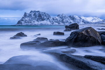 Langzeitbelichtung ds Meeresbrandung am Uttakleiv Beach, schneebedeckte Berge im Hintergrund