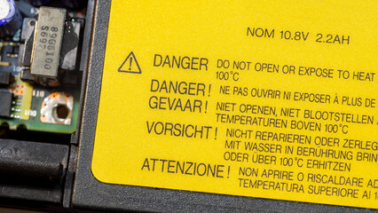 A yellow danger label with heat exposure caution message in various languages and a exclamation mark triangle on a generic electronic device laptop battery, object closeup detail User warnings concept