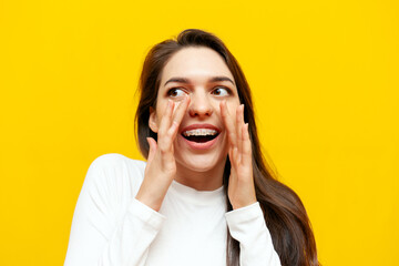 excited young woman with braces announces information and shouts over yellow isolated background,...