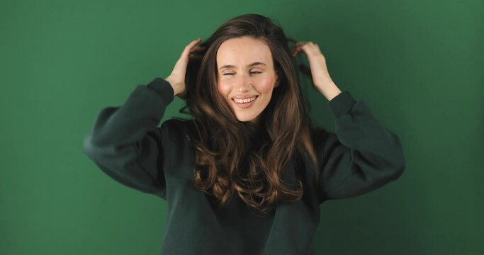 Cheerful happy joyful young woman wear green sweatshirt, brunette fixe hair. Smiling face. Happy woman fix hair, look funny and funky, bite lip isolated on green background.
