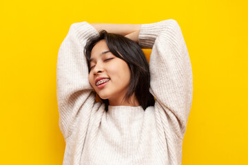young relaxed asian girl with braces resting with hands behind head on yellow isolated background,...