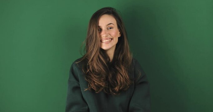 Woman look at camera see you, wave her hand, come to me. Hello, come to me! Portrait of friendly sociable woman in green sweatshirt gesturing hi, welcoming with raised hand stand on green background.