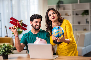 Indian young couple planning a vacation with laptop, piggy bank, toy plane and globe