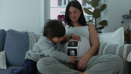 Child holding ultrasound image of baby brother next to pregnant mother seated on couch at home,...