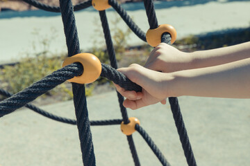 Hands of little preschooler and black rope as mesh for climbing on playground in park. Child development