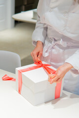 Uniformed baker clerk hands knotting a pink bow in a white gift box.