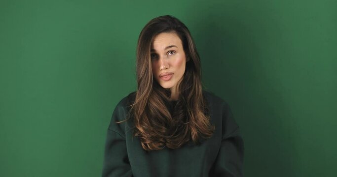 Upset curly brunette hair woman wear green pullover purses lips offendedly and looks from under forehead on green background. Sad tired woman looks with poker face at camera purses lip.