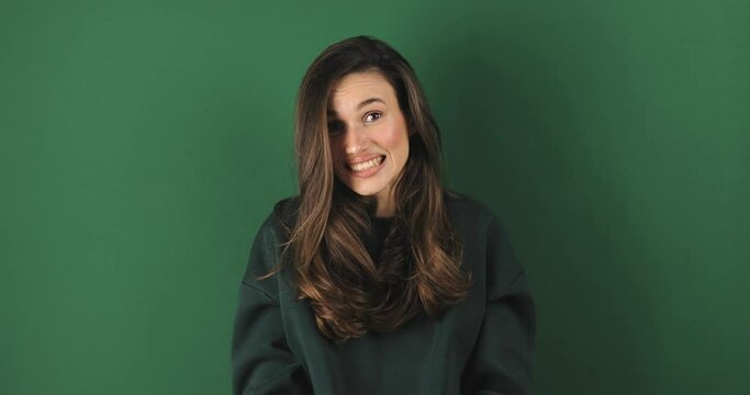 Shocked smiling shy charming confused young woman in green pullover posing isolated on green background studio. Girl looking at camera smile cover mouth with hand say oops.