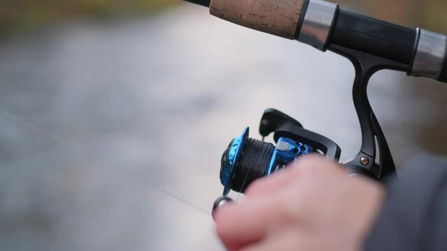 Zoom-in image of a fishing rod reel being turned by a human hand against a blurred river background