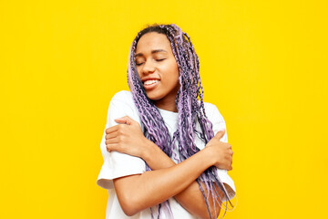 young african american woman with dreadlocks hugs herself over yellow isolated background, girl...