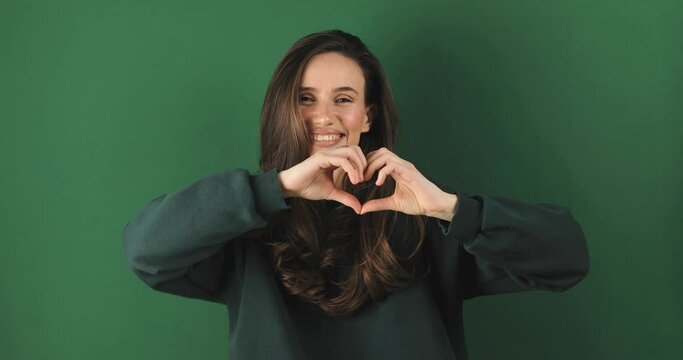 Happy attractive brunette woman look camera showing shape heart with hands, girl wear green sweatshirt isolated on green background. Love, romantic concept.