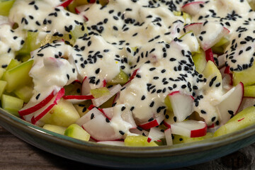 Red radish and cucumber salad with yogurt dressing and nigella seeds in a bowl standing on rustic wooden boards. 