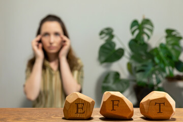 Letters EFT written on wooden blocks. Female tapping side of the eye (SE) meridian point in blurred background. Emotion-focused therapy treatment concept. - obrazy, fototapety, plakaty