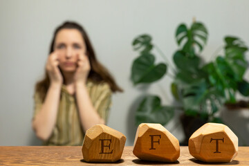Letters EFT written on wooden blocks. Female tapping under eye (UE) meridian point in blurred background. Emotion-focused therapy treatment concept.