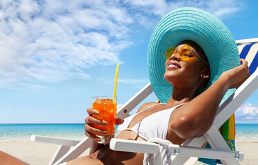 Happy woman is sunbathing on a beach deck chair, wearing sun hat and sunglasses, drinking a orange juice on a sunny day by the seaside, concept of a summer beach holiday, booking travel and resort