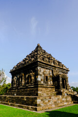 Obraz premium A large stone temple with a pyramidal roof, surrounded by lush green grass. The temple appears to be an ancient Hindu or Buddhist structure with architectural details.