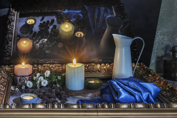 A still life with a blue satin fabric next to several lit candles, flowers surrounded by a golden...