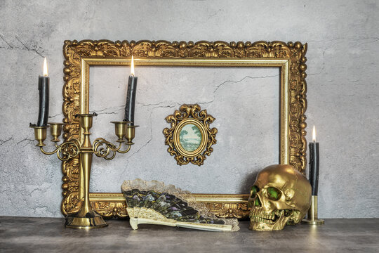 A decorative still life with a golden frame next to a golden skull next to several candelabras and a fan decorated with flowers