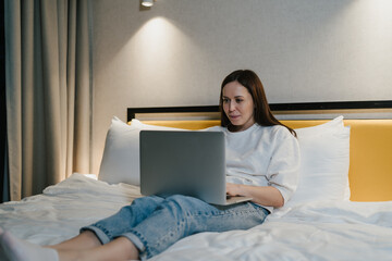 Relaxed woman engaged in using her laptop while comfortably reclining on bed, telecommuting or enjoying leisure time.