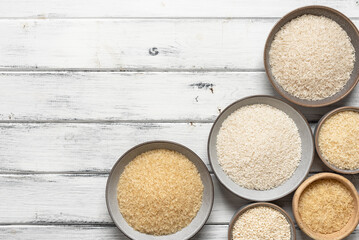 Assortment of different varieties of dry rice in bowls on a white wooden background. Top view, copy space.