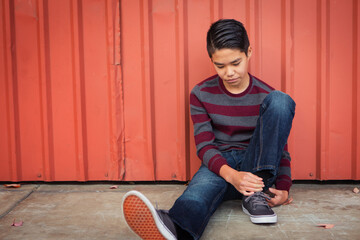 Lonely, sad Asian boy sitting back against a metal wall while gazing down.