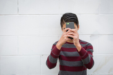 Teenage asian boy standing alone against an outdoor white background, holding his smartphone up to...