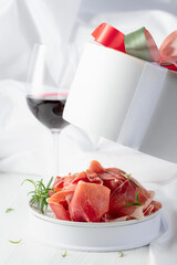 Prosciutto with rosemary in gift box and glass of red wine on a white wooden table.