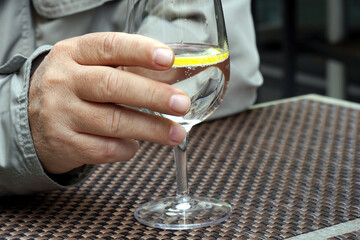 A male hand holding a wine glass with water and a slice of lemon in café sitting outside. 