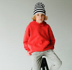 Young Boy With Curly Hair Poses in Red Hoodie and Striped Hat
