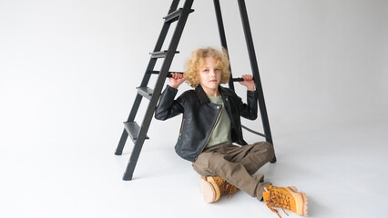 Young Boy With Curly Hair Sitting Under Black Ladder in Studio Setting