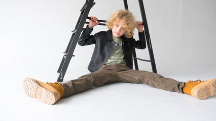 A young guy with curly hair is sitting on the floor with his legs spread wide against the background of the back stairs, fashion and style.