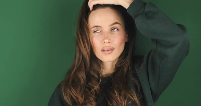 Cheerful happy joyful young woman wear green sweatshirt, brunette fixe hair. Smiling face. Happy woman fix hair, look funny and funky, bite lip isolated on green background.