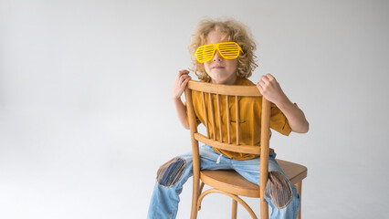 A curly-haired red-haired boy in stylish yellow glasses is sitting on a chair looking at the front.