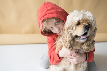 A happy child in a red sweatshirt with a hood on his head plays and hugs a Cocker spaniel dog.
