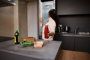 Young woman pulling out a glass bottle with red juice of tomatoes from the refrigerator. Housewife...