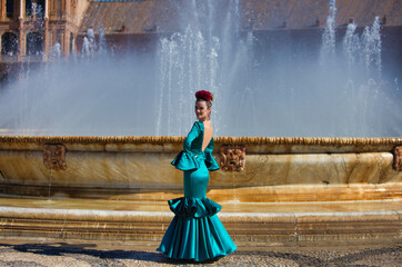 Beautiful young woman in a green frilly suit with a flower on her head. The woman is dancing flamenco and is in the most famous square in seville, spain, in front of its central fountain.