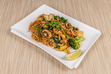 A plate of Chinese food, thin lo mein noodles with chicken meat and and broccoli and vegetables, and other dishes in the background, on a wooden tabl