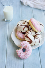 Frosted vanilla donuts with chocolate swilrs and strawberry pink doughnuts with coconut flakes. Bite missing. Glass of milk in background. Selective focus with blurred foreground and background.