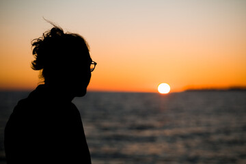 Un ragazzo con i capelli lunghi e gli occhiali guarda il tramonto su una spiaggia a Marina di Ragusa