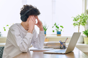 Sad upset young guy in headphones sitting at home at desk with computer