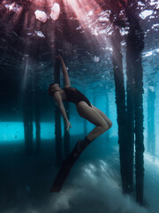 Freediver woman underwater under the pier in blue sea with sun rays. - 780792668