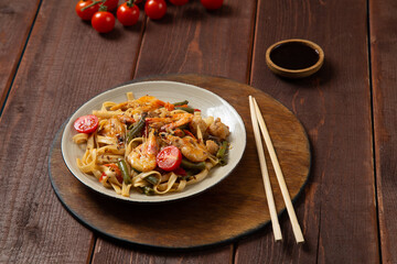 udon shrimp in pad thai sauce on a wooden stand next to cherry tomatoes, soy sauce and chopsticks
