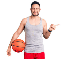 Young handsome man holding basketball ball smiling happy pointing with hand and finger to the side