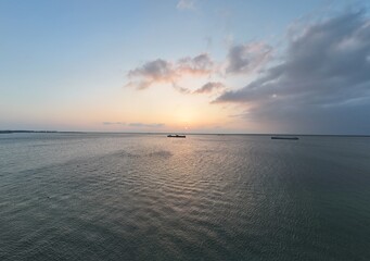 Serene seascape at sunrise with calm waters and a clear sky, featuring distant boats on the horizon under the soft glow of the morning sun.