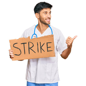Young handsome man wearing doctor uniform holding strike banner cardboard pointing thumb up to the side smiling happy with open mouth