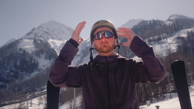 Close-up: A male skier puts on sunglasses. A guy gets dressed on a ski slope in snowy mountains. The snowboarding resort in winter. Man face detail adjusting protective helmet preparing for skiing.